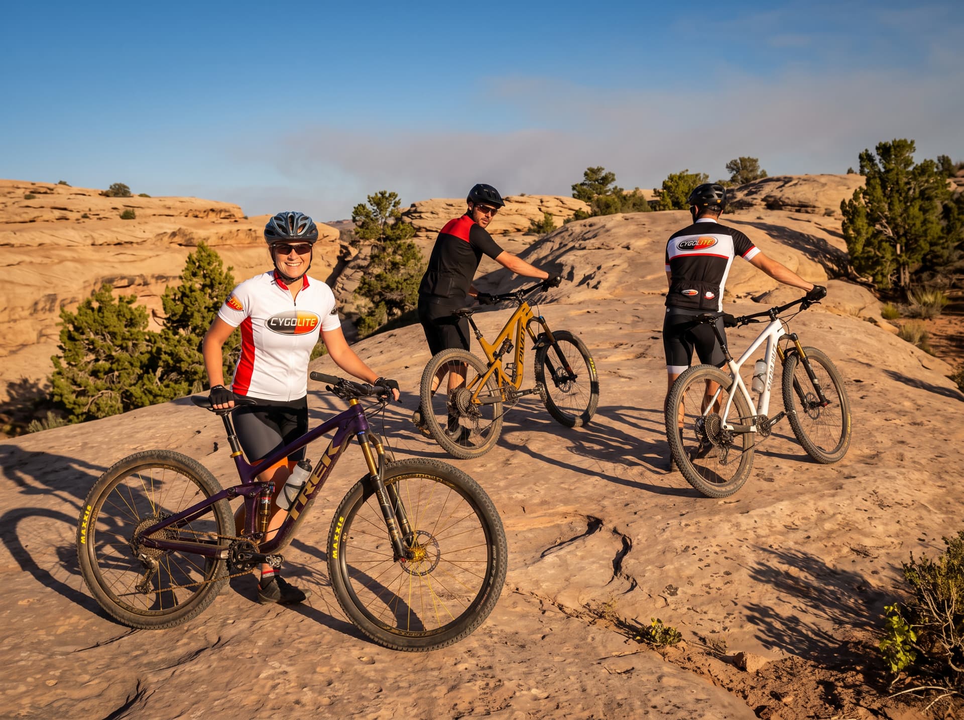 Three cyclists in Cygolite jerseys with mountain bikes staging on desert slickrock at golden hour